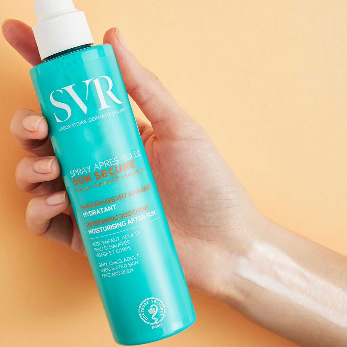 Türkisblaue SVR SUN SECURE After-Sun-Sprühflasche in der Hand mit weißem Sprühkopf vor beigefarbenem Hintergrund.