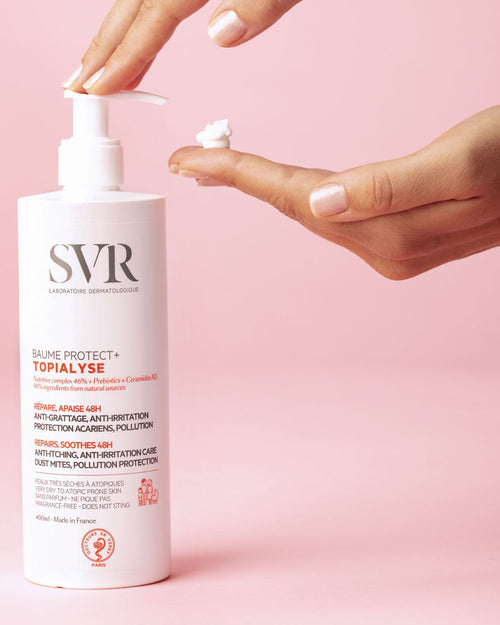 Weiße SVR Topialyse Baume Protect+ Pumpflasche mit weißer Creme auf einer Hand vor einem hellrosa Hintergrund.