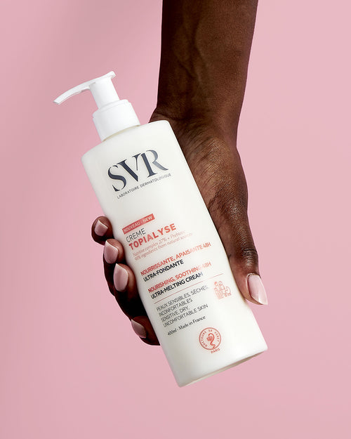 Hand hält weiße 400ml SVR TOPIALYSE Creme Pumpflasche vor einem hellrosa Hintergrund.