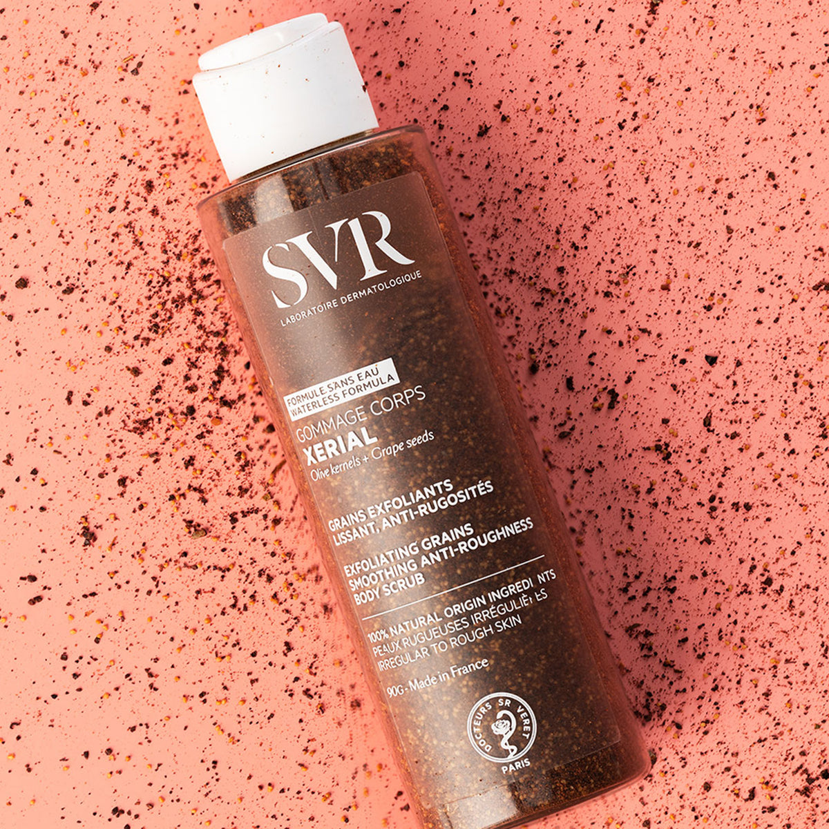 SVR Xerial Gommage Körperpeeling in transparenter Flasche mit Oliven- und Traubenkernen auf einem korallenfarbenen Untergrund.