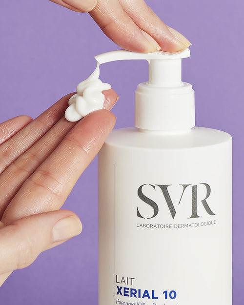 Weiße SVR XERIAL 10 Lait Pumpflasche mit cremiger Textur auf der Hand vor lila Hintergrund.