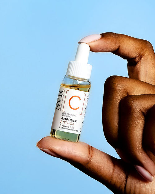 Hand hält eine kleine Glasflasche SVR Ampoule Anti-Ox Vitamin C Serum mit weißem Pipettenverschluss vor blauem Hintergrund.