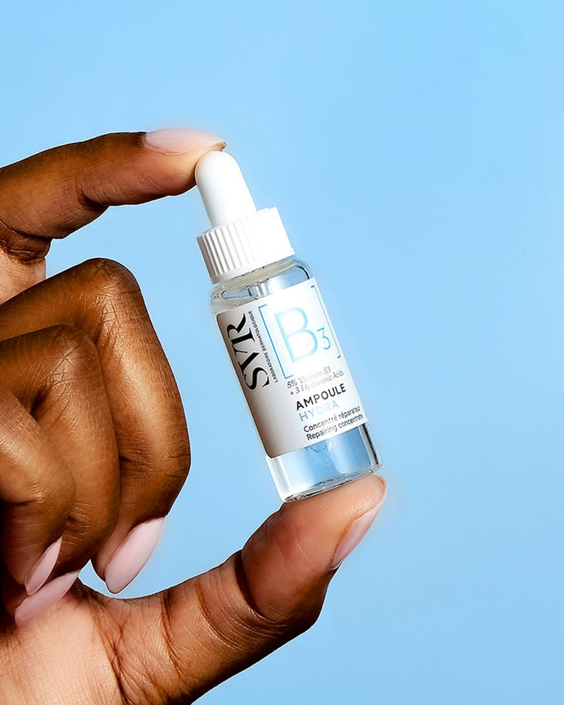 Hand hält eine kleine, transparente SVR B3 Ampoule Hydra Glasflasche mit weißem Pipettenverschluss vor blauem Hintergrund.