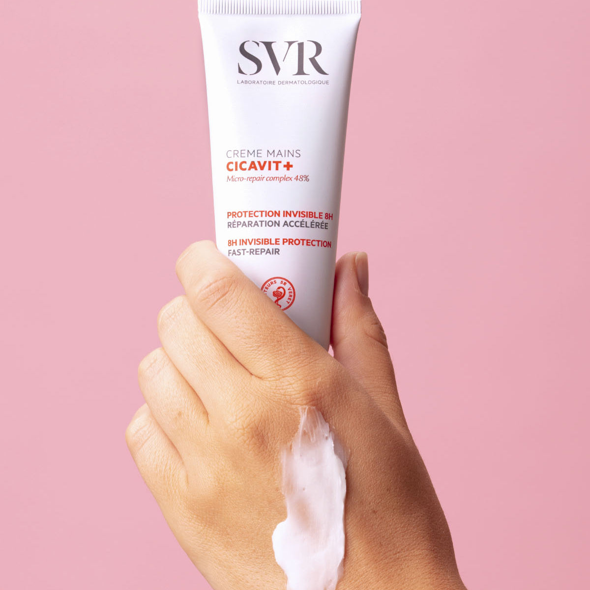 Hand hält eine weiße SVR Cicavit+ Handcremetube mit roter Schrift vor einem pastellrosa Hintergrund mit Creme auf dem Handrücken.