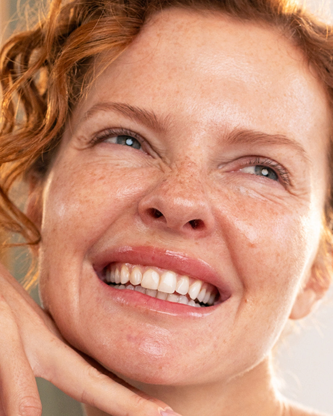 Close-up of a smiling woman with red curly hair and freckled skin, showing a radiant and healthy complexion.