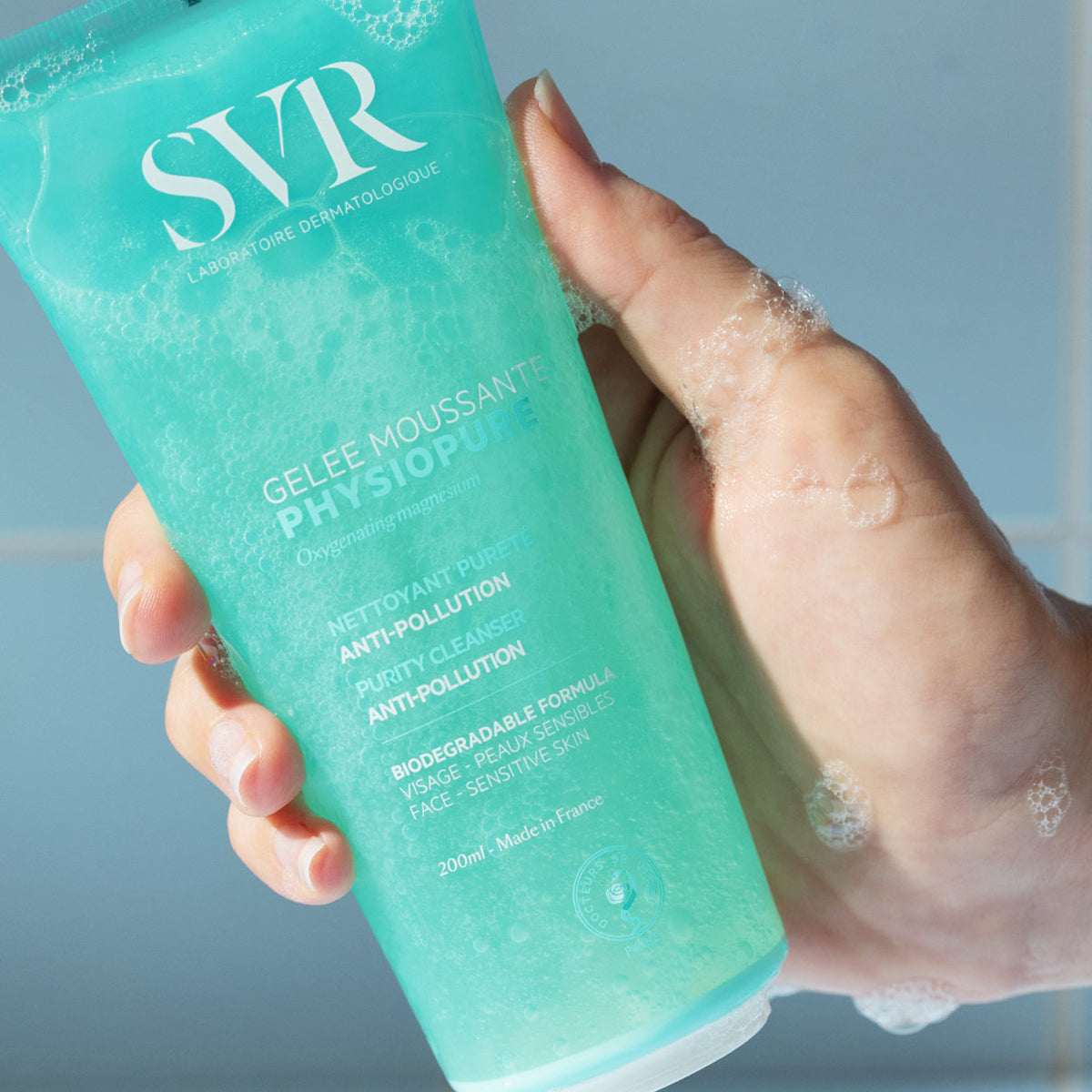 Hand hält eine türkisfarbene SVR PHYSIOPURE Gelée Moussante Tube mit schäumender Textur vor einem hellen Hintergrund.