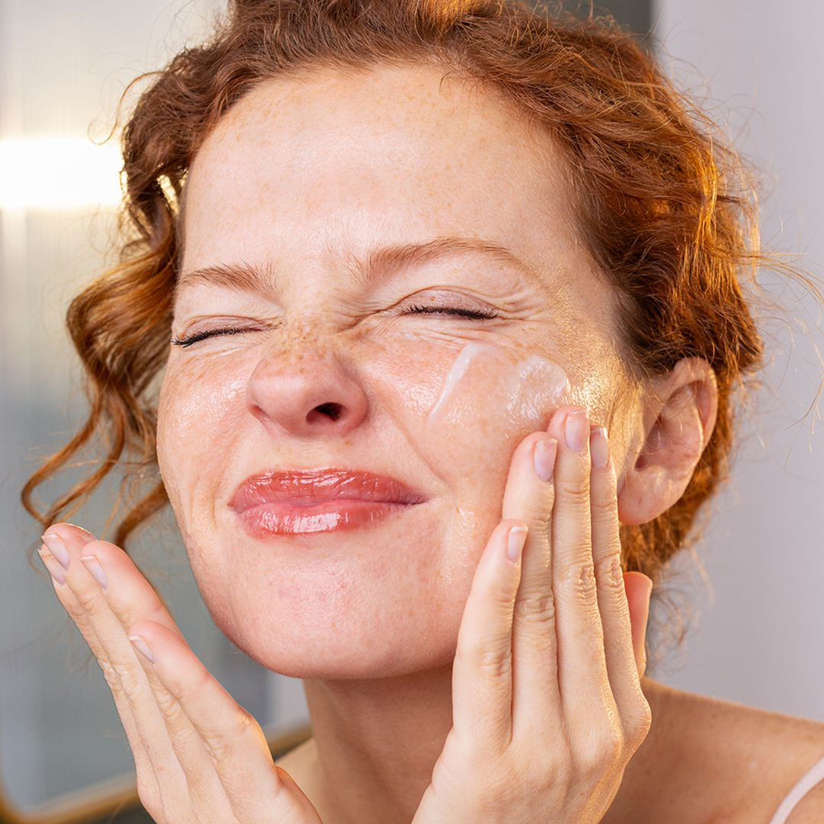 Frau mit lockigem Haar trägt SVR Sensifine Reinigungsbalsam auf ihr Gesicht auf, um Make-up sanft zu entfernen.