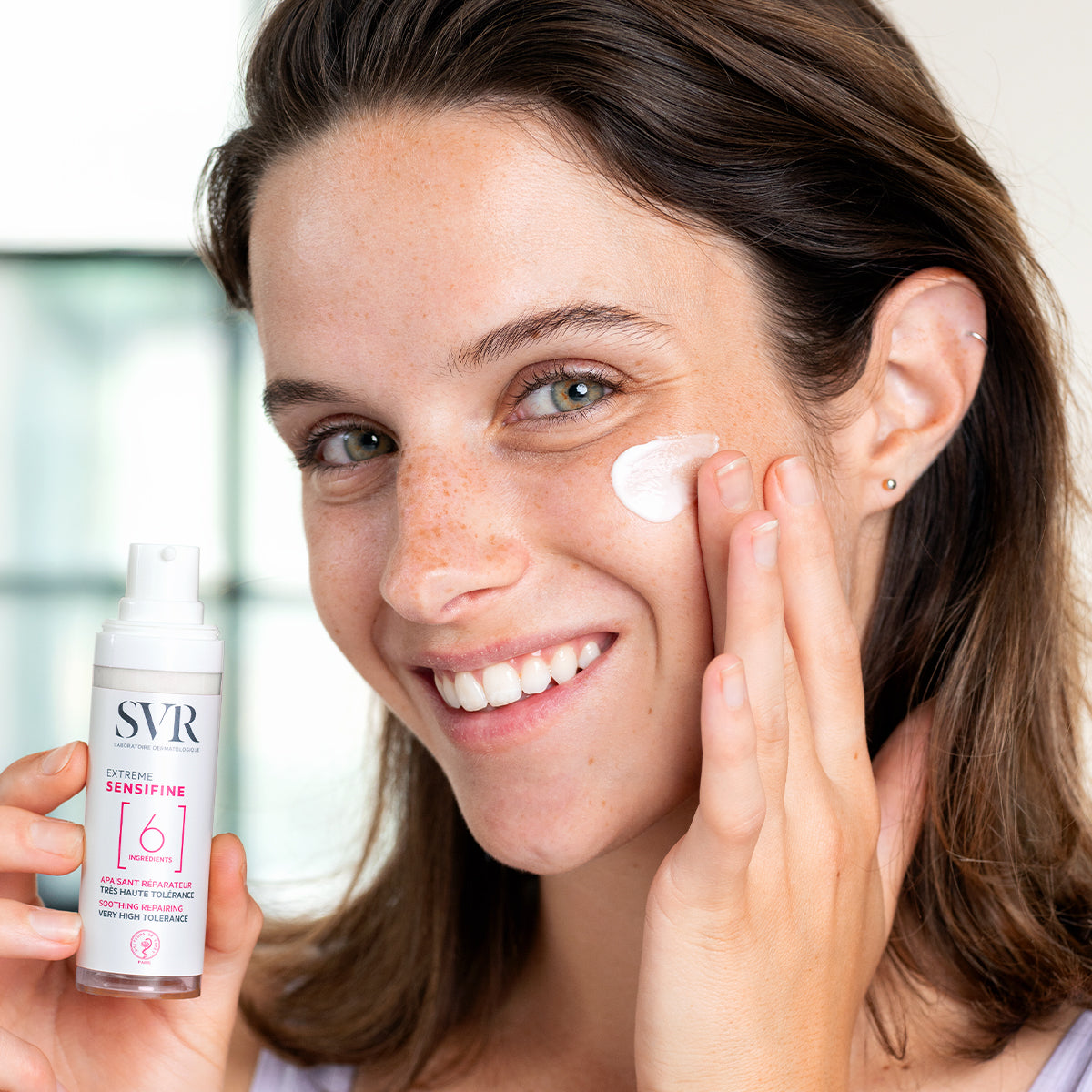 Woman applying white SVR Sensifine Extreme soothing cream from a pump bottle onto her cheek.
