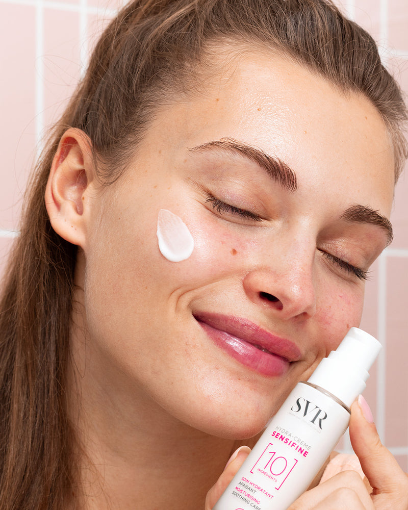 Woman applying white SVR SENSIFINE Hydra-Crème from a pump bottle to her cheek against a pink tiled background.