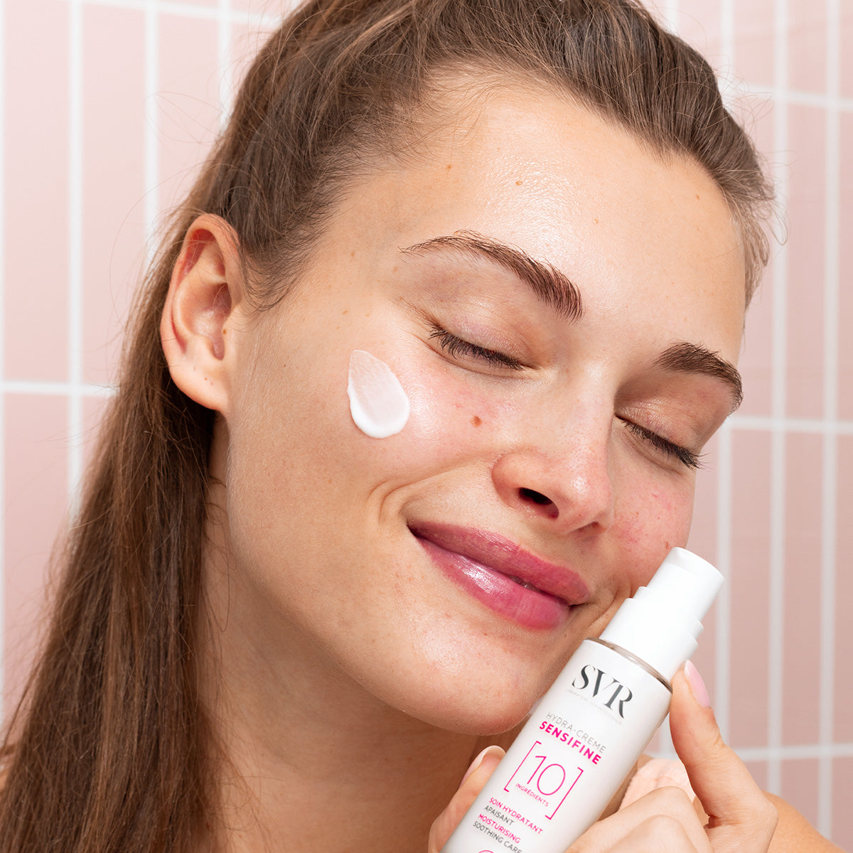 Woman applying white SVR SENSIFINE Hydra-Crème from a pump bottle to her cheek against a pink tiled background.