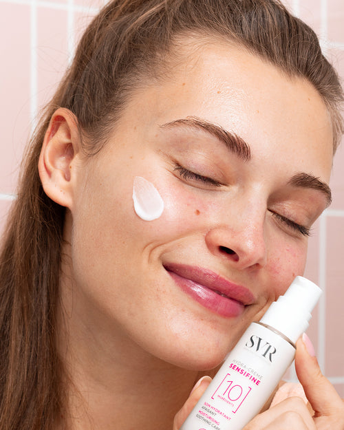 Woman applying white SVR SENSIFINE Hydra-Crème from a pump bottle to her cheek against a pink tiled background.