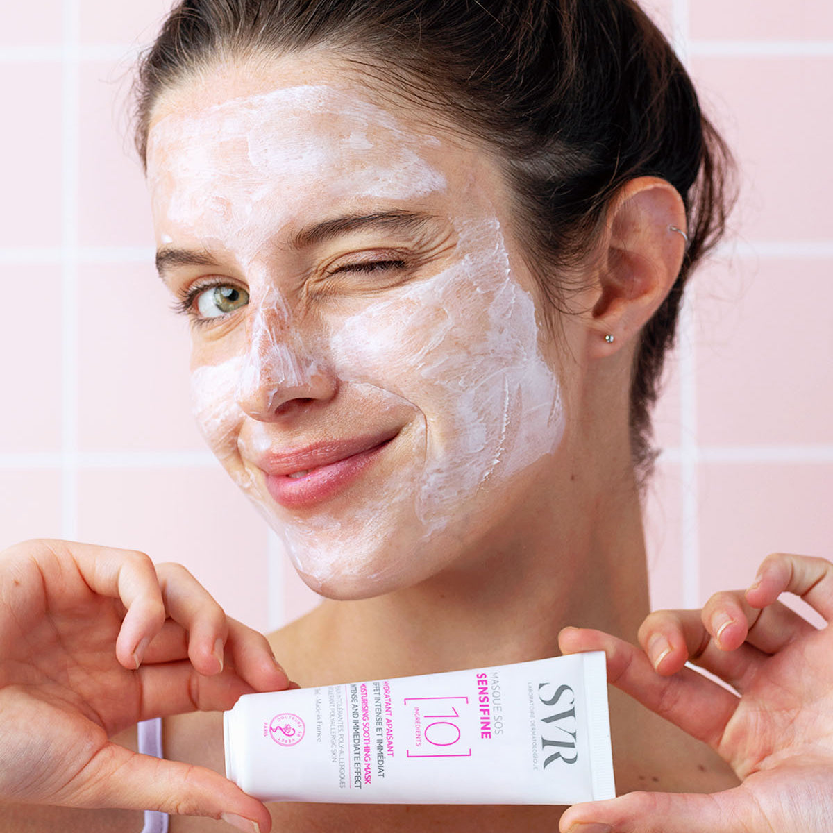 Frau mit weißer SVR SENSIFINE Masque SOS Gesichtsmaske, die eine weiße Tube des Produkts vor rosa Fliesen hält.
