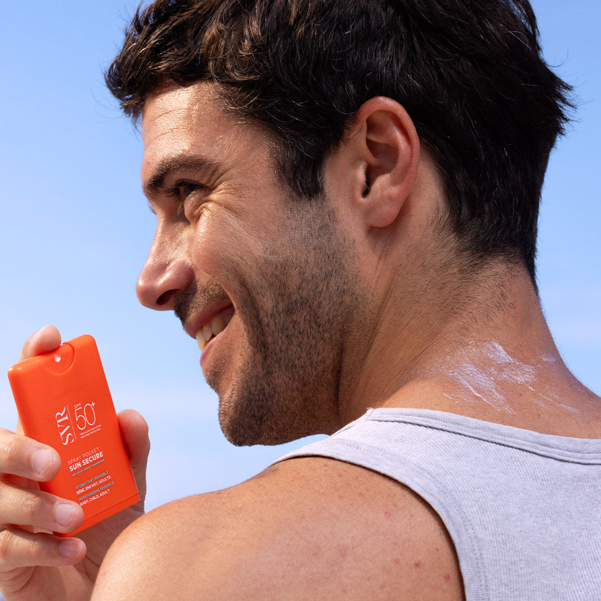 Mann hält eine orangefarbene SVR Sun Secure SPF50+ Taschenspray-Flasche vor blauem Himmel.
