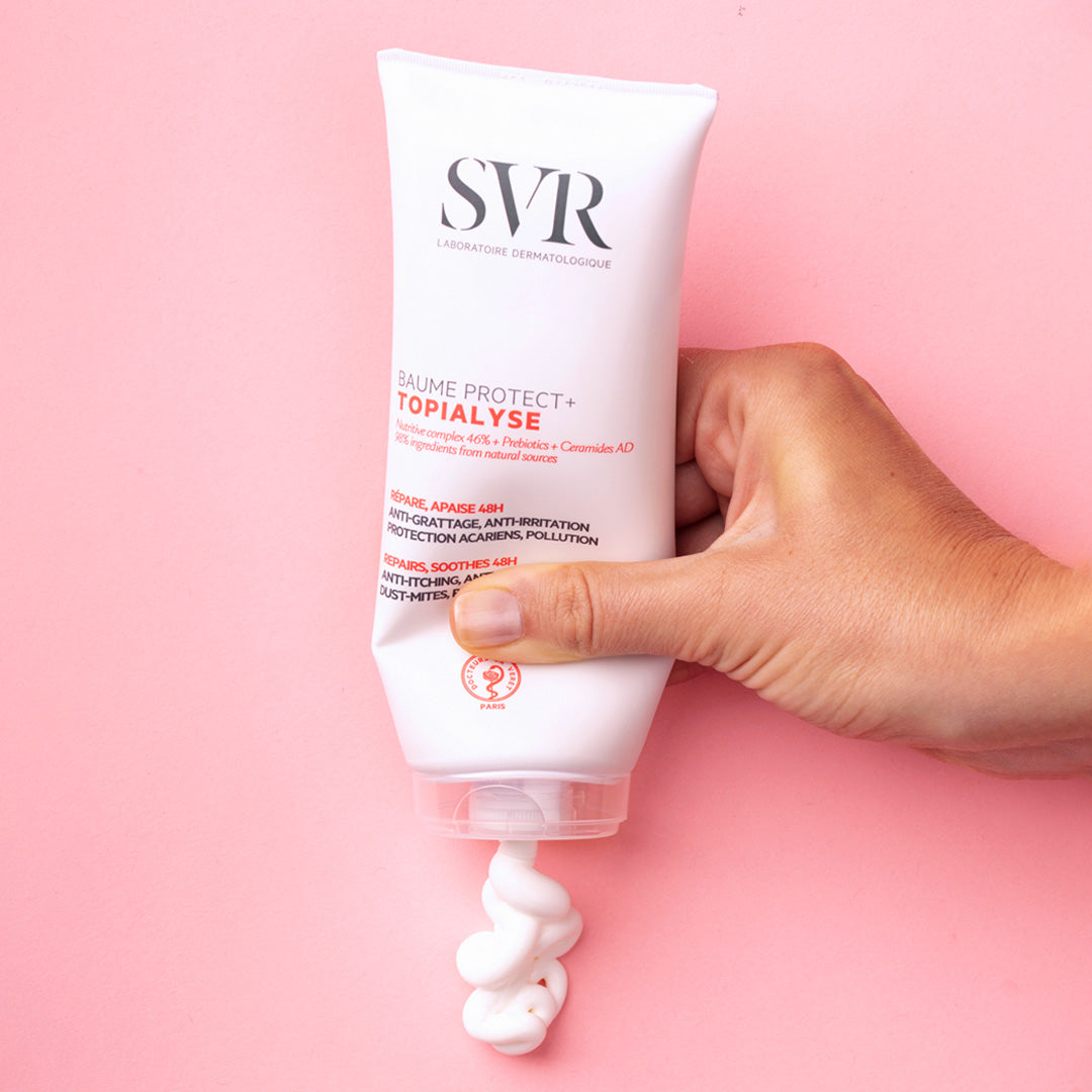 Hand hält eine weiße Tube SVR TOPIALYSE Baume Protect+ Creme, aus der eine reichhaltige, weiße Textur auf rosa Hintergrund austritt.