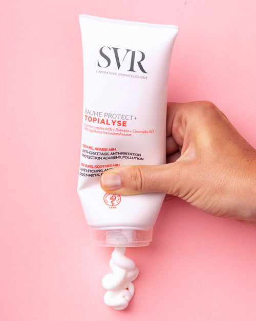 Hand hält eine weiße Tube SVR TOPIALYSE Baume Protect+ Creme, aus der eine reichhaltige, weiße Textur auf rosa Hintergrund austritt.