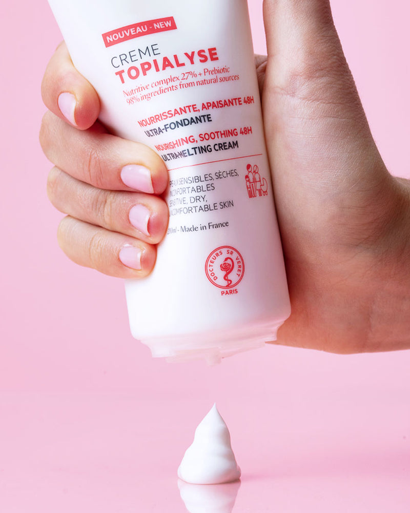 Hand hält weiße TOPIALYSE Crème Tube vor rosa Hintergrund, aus der eine Portion reichhaltige, weiße Pflegecreme herausgedrückt wird.