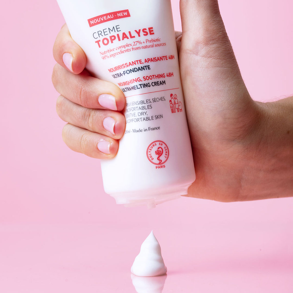 Hand hält weiße TOPIALYSE Crème Tube vor rosa Hintergrund, aus der eine Portion reichhaltige, weiße Pflegecreme herausgedrückt wird.