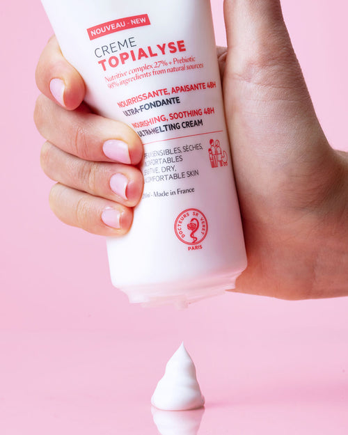 Hand hält weiße TOPIALYSE Crème Tube vor rosa Hintergrund, aus der eine Portion reichhaltige, weiße Pflegecreme herausgedrückt wird.