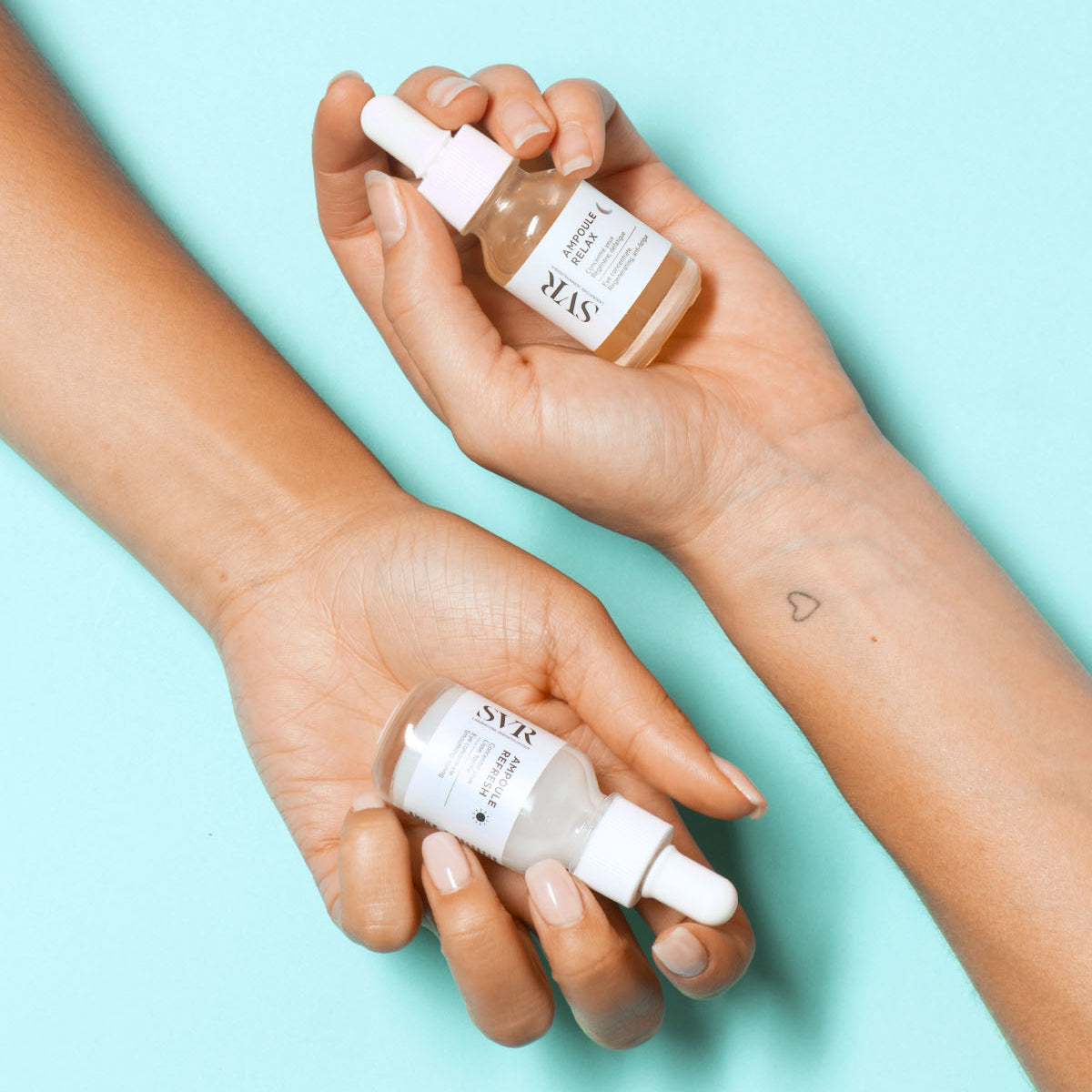 Zwei Hände halten SVR Ampoule Relax und Refresh Augenserum in Glasflaschen mit weißer Pipette auf hellblauem Hintergrund.