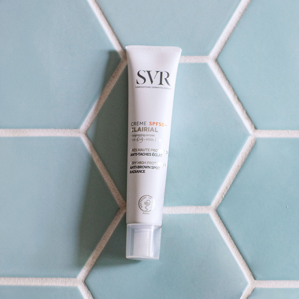 Weiße SVR Clairial Crème SPF50+ Tube auf hellblauen, sechseckigen Fliesen mit weißer Fuge.