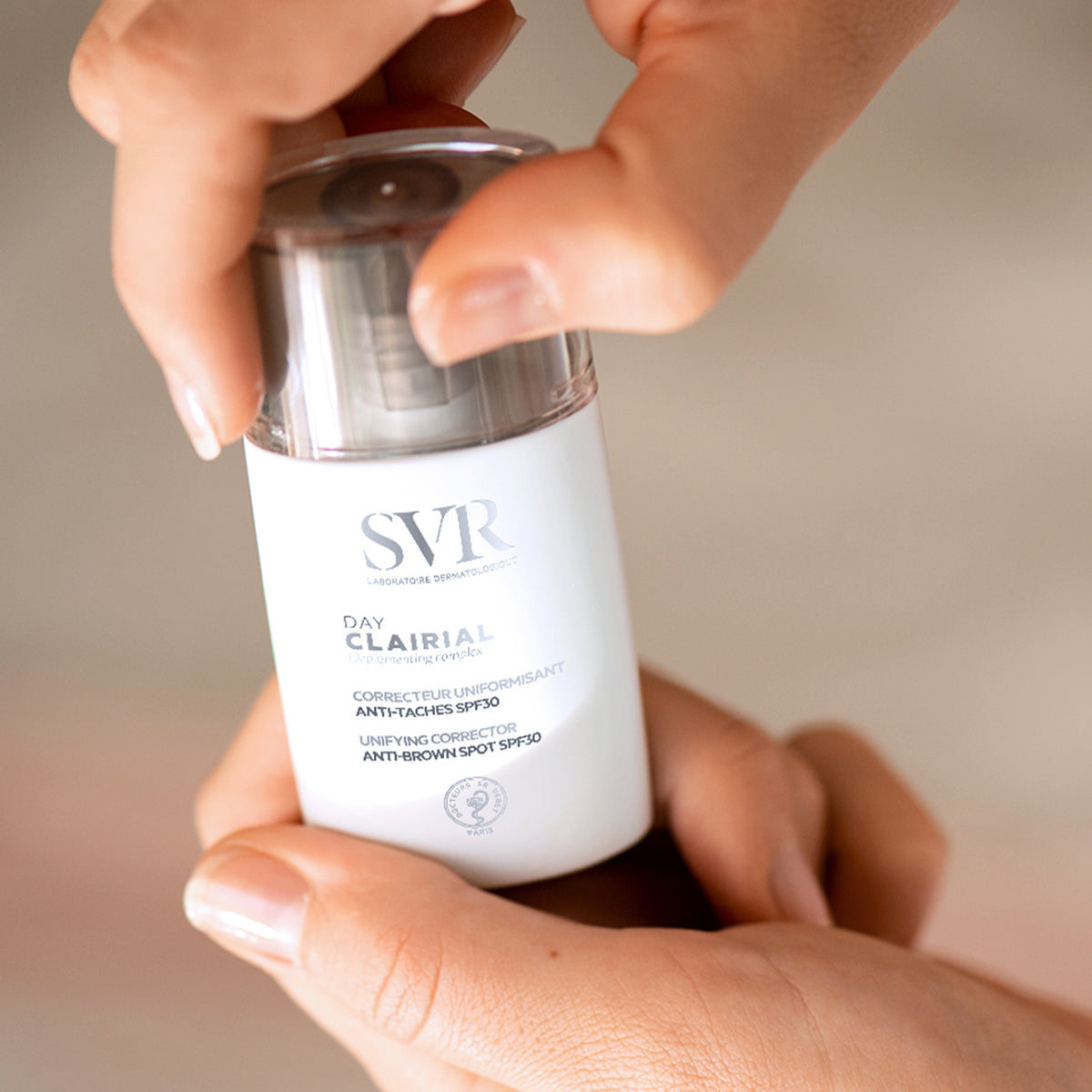 Hände halten eine weiße SVR Clairial Day Flasche mit transparentem Deckel und SPF30 Aufdruck zur Pigmentflecken-Korrektur.