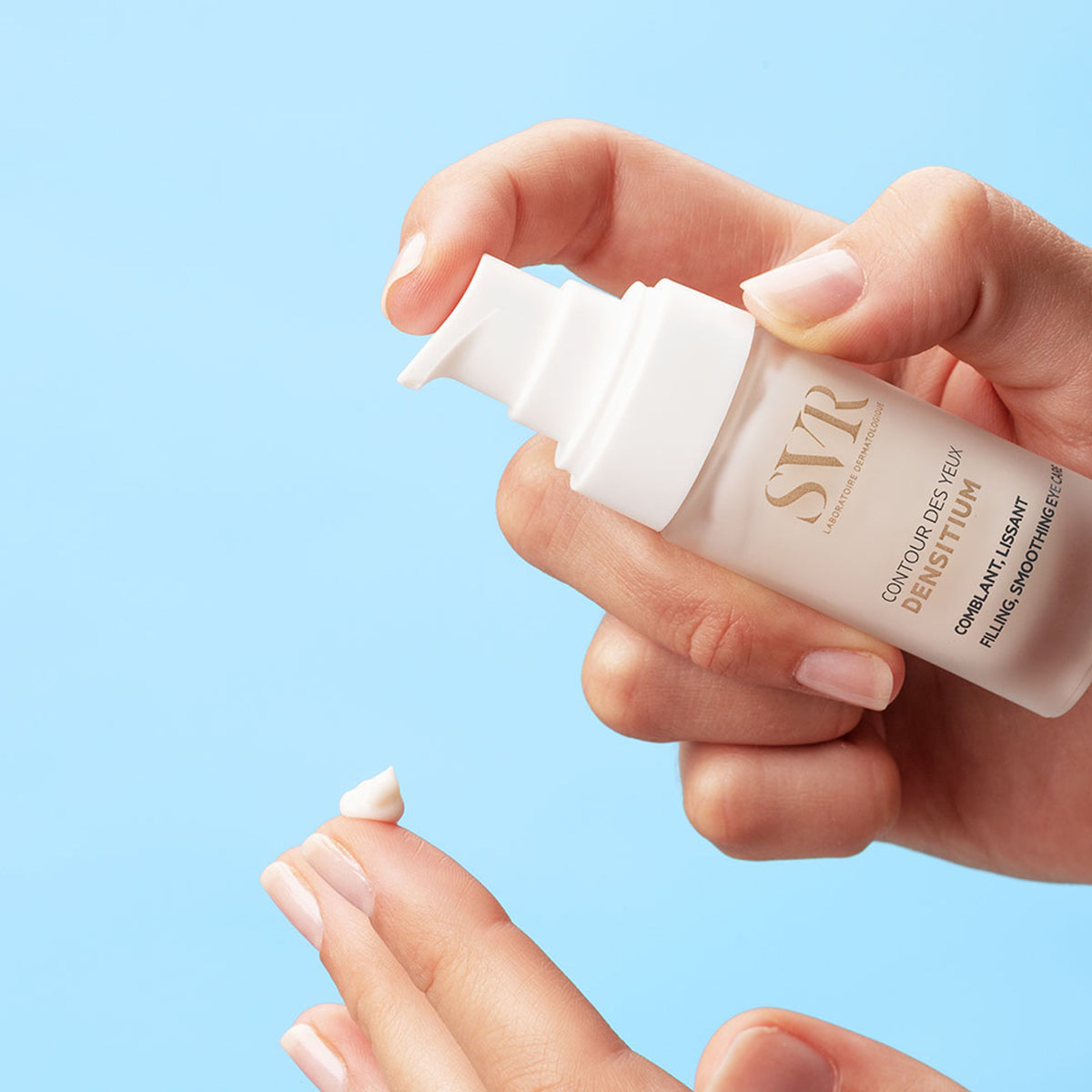 Hand drückt SVR DENSITIUM Contour des Yeux Augencreme aus dem weißen Pumpspender auf den Finger vor blauem Hintergrund.