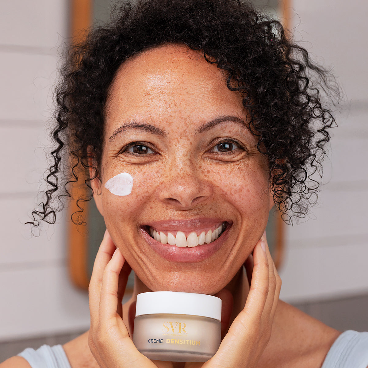 Smiling woman holding a jar of SVR DENSITIUM Crème with a dab of white face cream on her cheek.