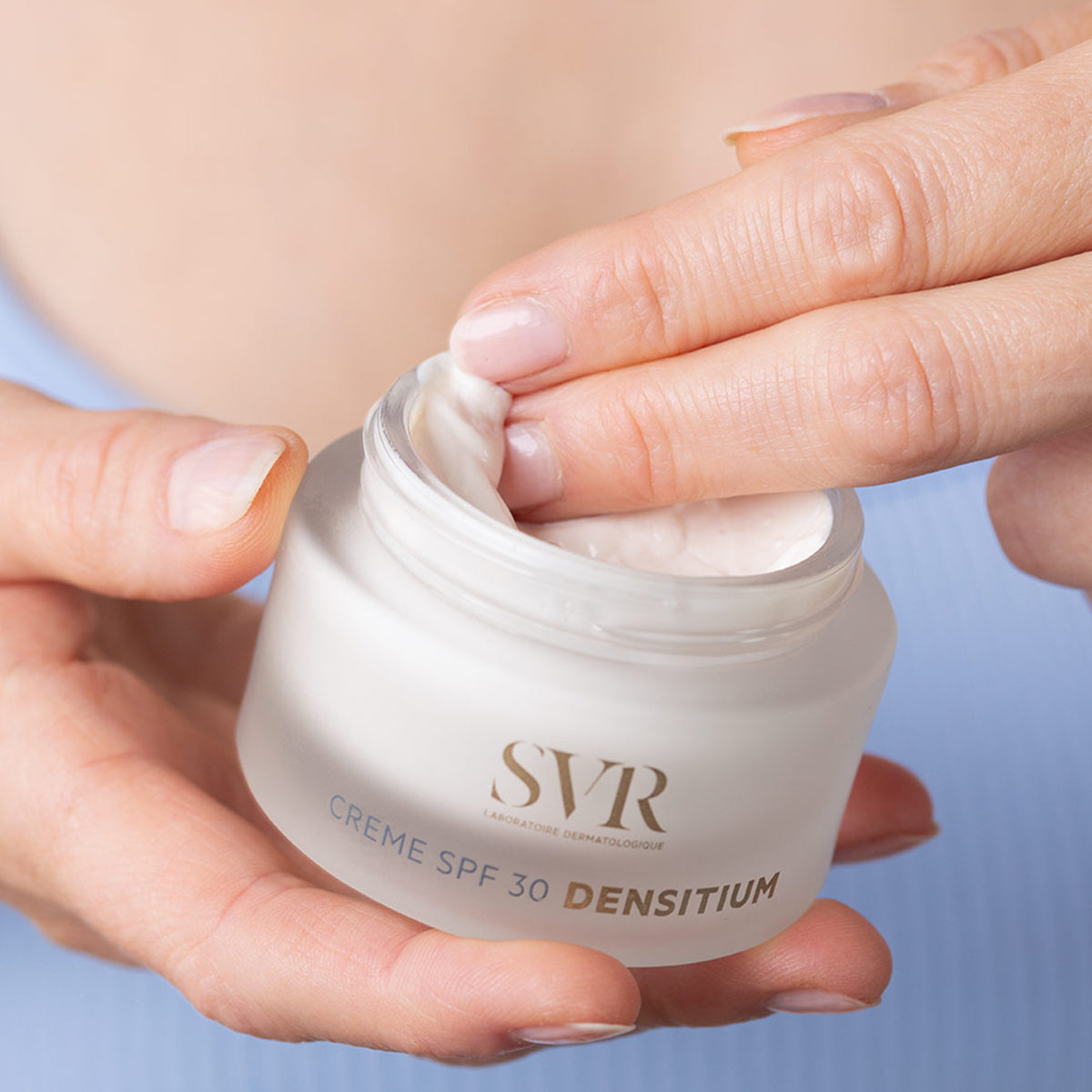 Hands holding a frosted glass jar of SVR DENSITIUM Crème SPF30 with a finger scooping the rich white face cream.