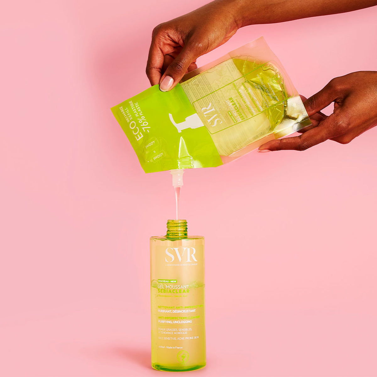 Hände füllen SVR SEBIACLEAR Gel Moussant Nachfüllbeutel in eine grüne, transparente 400ml-Flasche vor rosa Hintergrund.