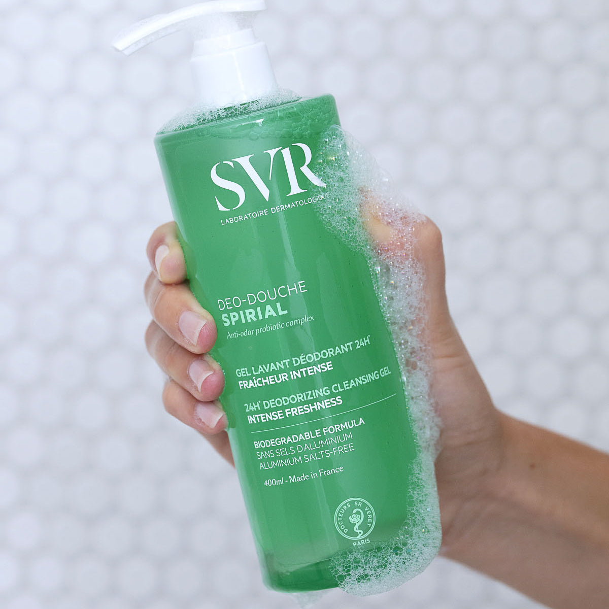 Grüne SVR Spirial Déo-Douche Pumpflasche mit Schaum in der Hand vor einem weißen Fliesen-Hintergrund.