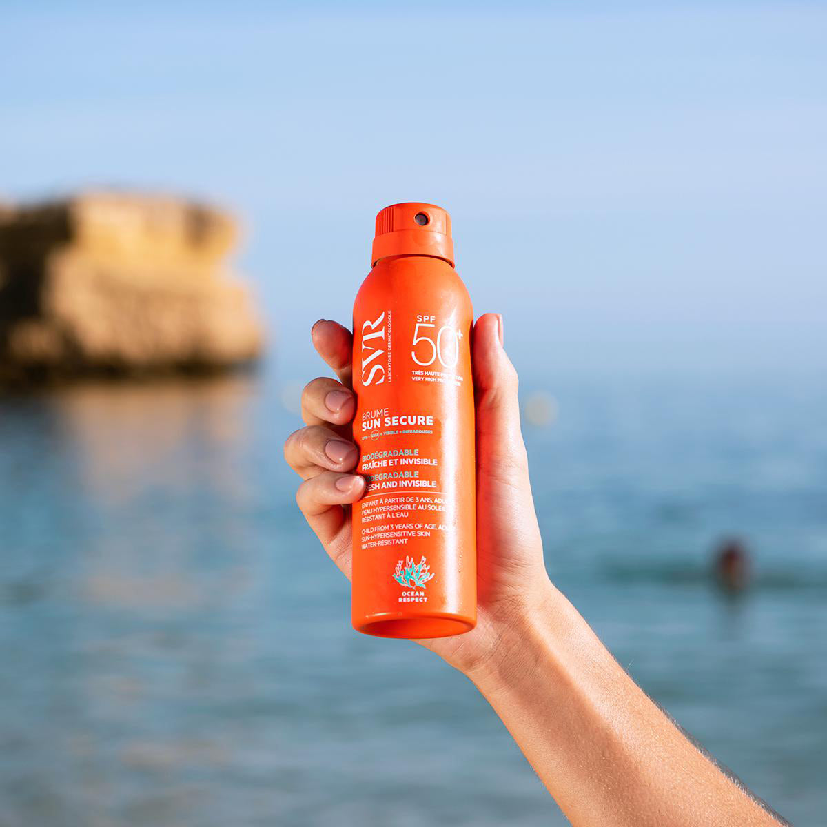 Hand hält orangefarbene SVR SUN SECURE Brume SPF50 Sonnenschutz-Spraydose vor einem Strandhintergrund.