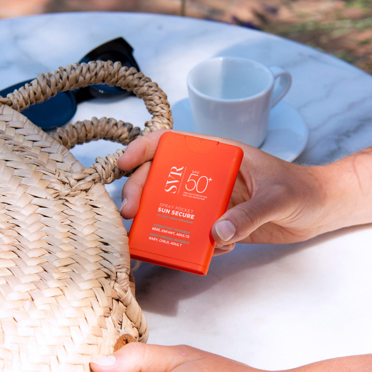 Hand hält orangefarbenes SVR SUN SECURE Spray Pocket SPF50+ vor einer Strohtasche und einem Kaffeetisch im Freien.