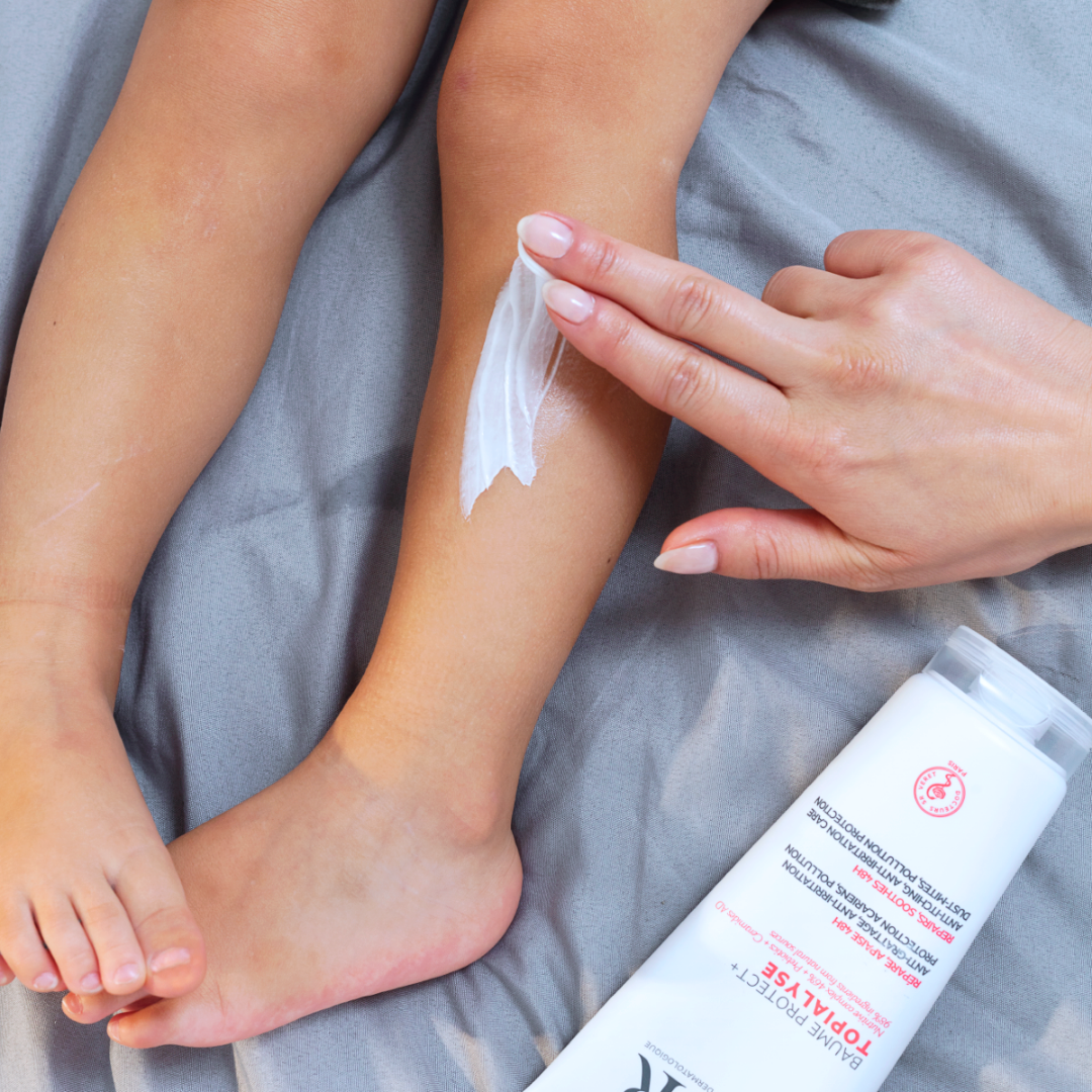 Hand trägt weiße TOPIALYSE Baume Protect+ Creme auf das Bein eines Kindes auf, vor einem grauen Stoffhintergrund.