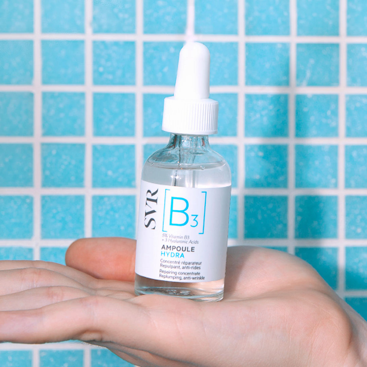 SVR B3 Ampoule Hydra Serum in einer Glasflasche mit weißem Pipettenaufsatz, gehalten in einer Hand vor blauen Mosaikfliesen.