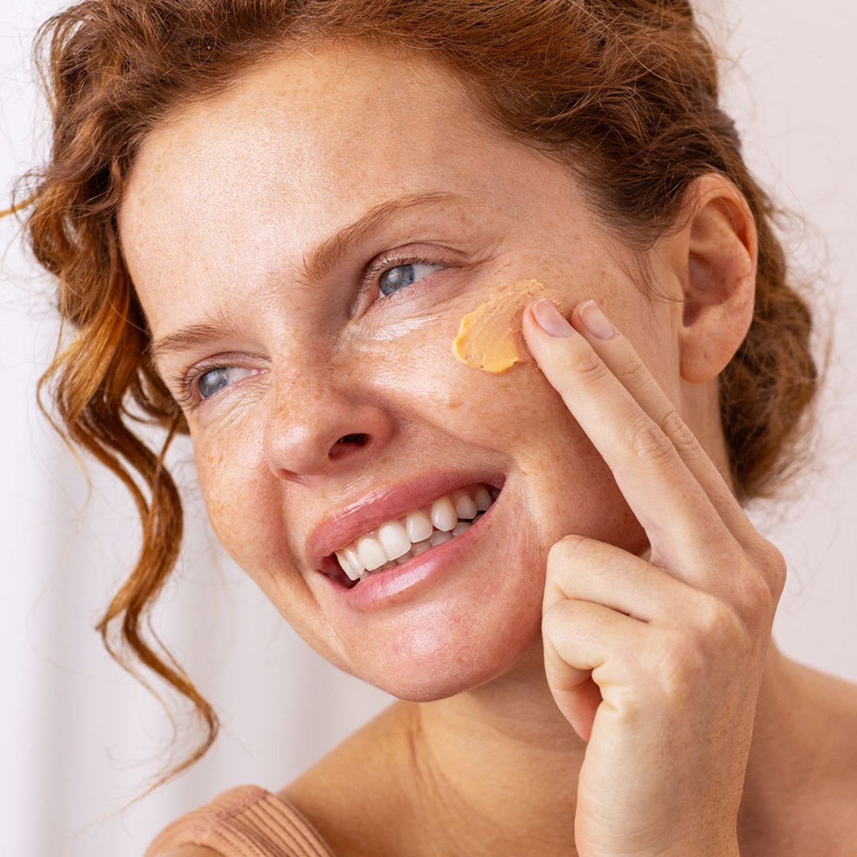 Frau mit rotem Haar trägt aprikosenfarbene Sun Secure Blur SPF50+ Mousse-Creme auf ihre Gesichtshaut auf.