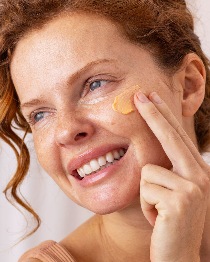 Frau mit rotem Haar trägt aprikosenfarbene SUN SECURE Blur SPF50+ Sonnencreme auf ihre Wange auf.