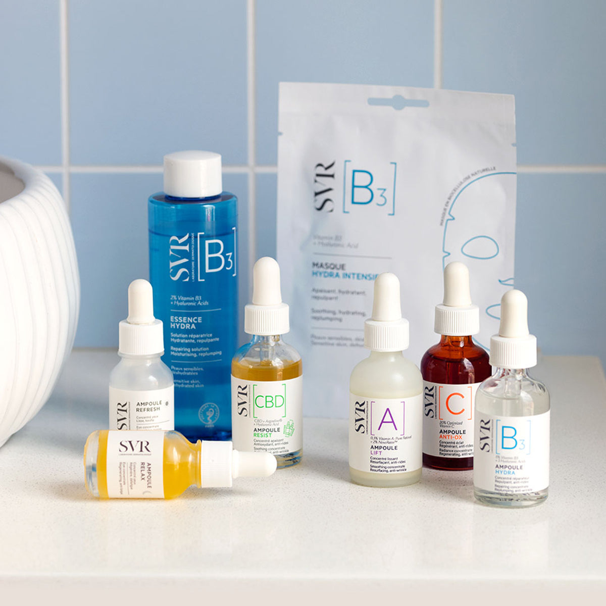 SVR Ampoule Relax und weitere SVR Hautpflege-Seren in Glasflaschen mit Pipette auf einer weißen Ablage vor blauen Fliesen.