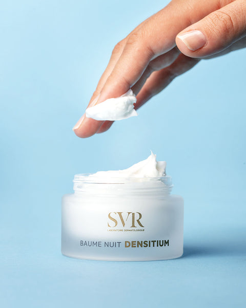 Finger picking up a dollop of white SVR Densitium night cream from an open frosted glass jar against a blue background.