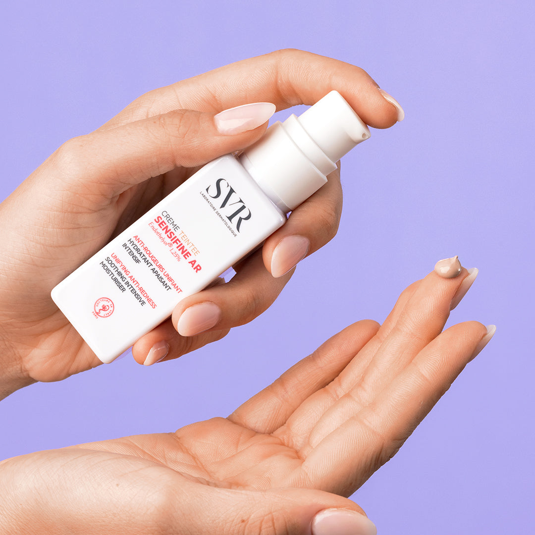 SVR Sensifine AR getönte Creme in weißem Pumpspender mit getöntem Produkt auf dem Finger vor lila Hintergrund.