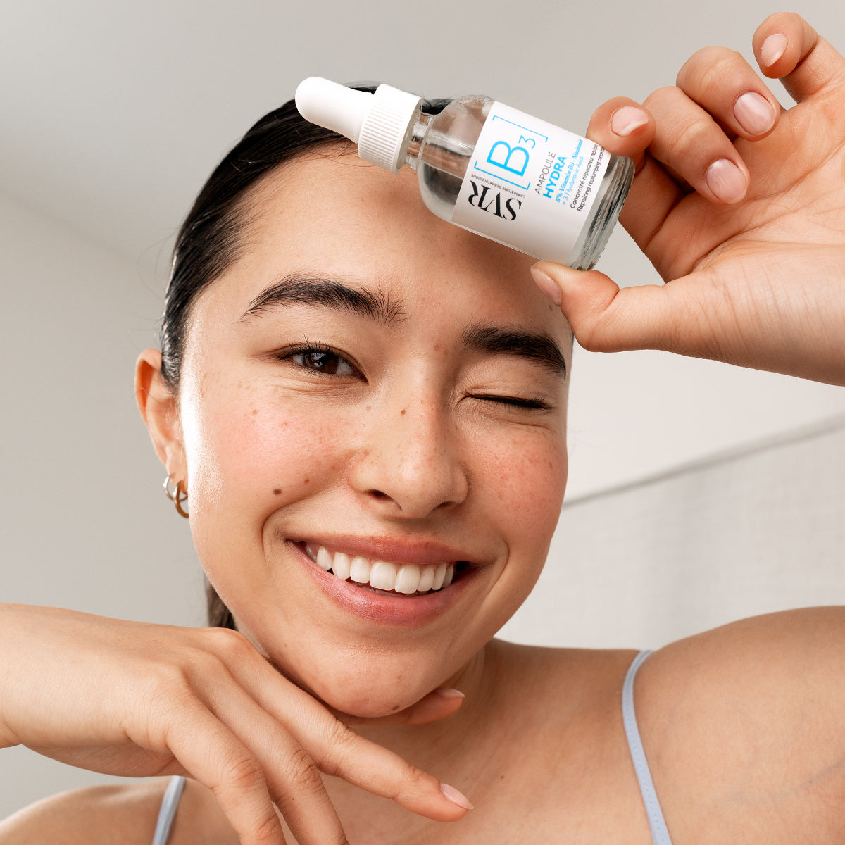 Lächelnde Frau hält ein SVR Ampoule Hydra Serum mit Vitamin B3 in einer transparenten Glasflasche mit weißem Pipettenverschluss.