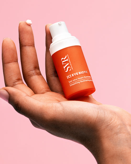 SVR C-EYE Biotic smoothing eye cream in an orange pump bottle held in a hand with a dab of white cream on a fingertip.