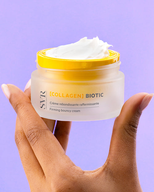 Hand holding a frosted glass jar of SVR Collagen Biotic firming cream with a bright yellow lid and white textured cream.