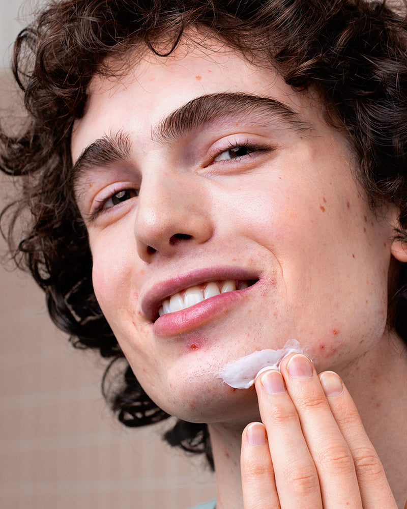 Young person with curly hair applying white CICAVIT+ Crème HPPI to a blemish on their chin in a bathroom setting.