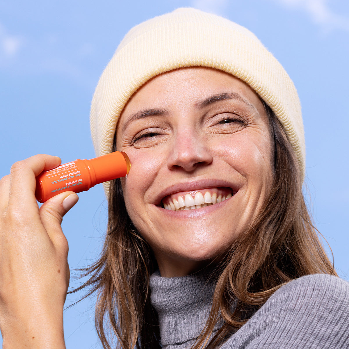 Lächelnde Frau trägt den orangefarbenen Sun Secure Easy Stick SPF50+ auf ihre Wange auf, vor hellblauem Hintergrund.