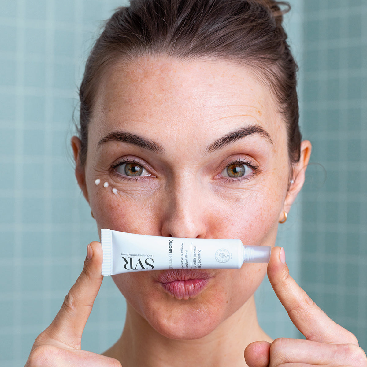 Frau hält weiße SVR [FILLER] BIOTIC Tube vor den Lippen mit Creme-Punkten unter den Augen für Augen- und Lippenpflege.