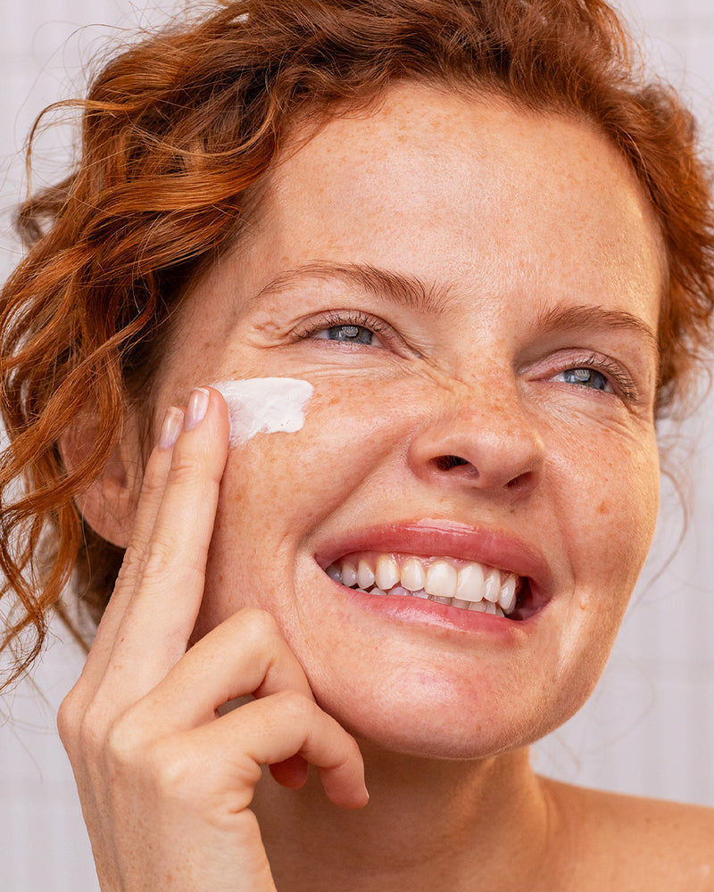 Frau mit rötlichem Haar trägt das SUN SECURE Fluide SPF50+ auf ihre Wange auf und lächelt strahlend in die Kamera.