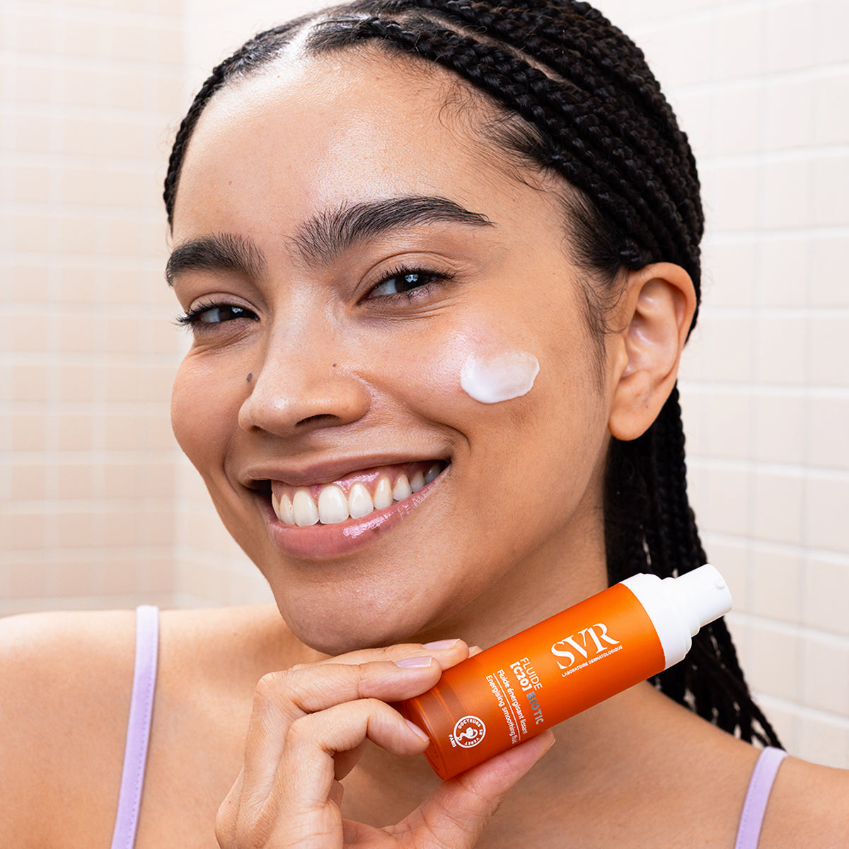 Smiling woman holding an orange SVR Fluide [C20] BIOTIC pump bottle with cream applied to her cheek.