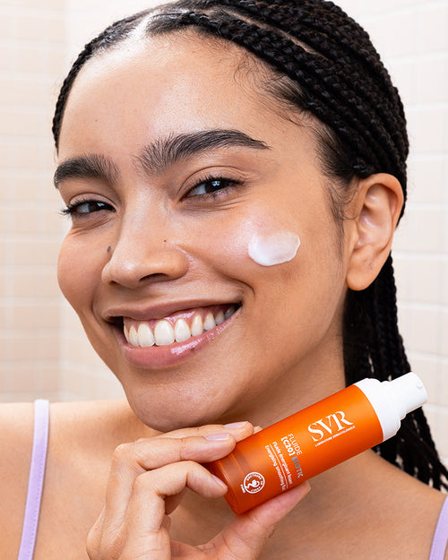 Smiling woman holding an orange SVR Fluide [C20] BIOTIC pump bottle with cream applied to her cheek.