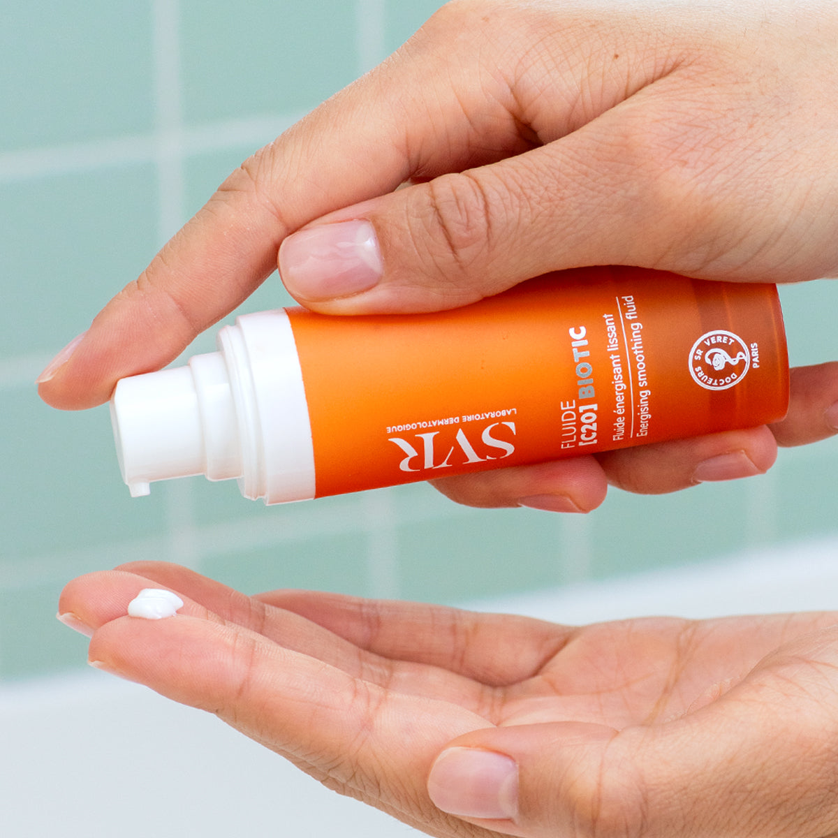 Hand holding an orange SVR Fluide [C20] BIOTIC pump bottle, dispensing a drop of white smoothing face cream into the palm.