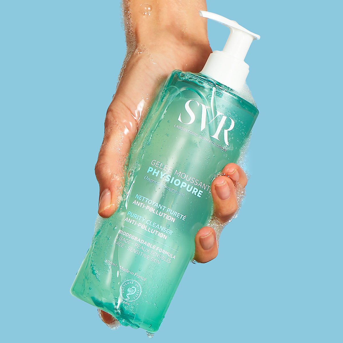 Hand hält eine türkisfarbene SVR PHYSIOPURE Gelée Moussante Flasche mit weißem Pumpspender und sanftem Reinigungsschaum.